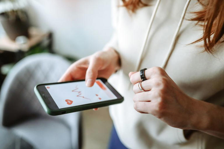 woman in a sporty hoodie with a smart ring and smartphone at home checks a heart rate and pulse monitoring app, technology in everyday life