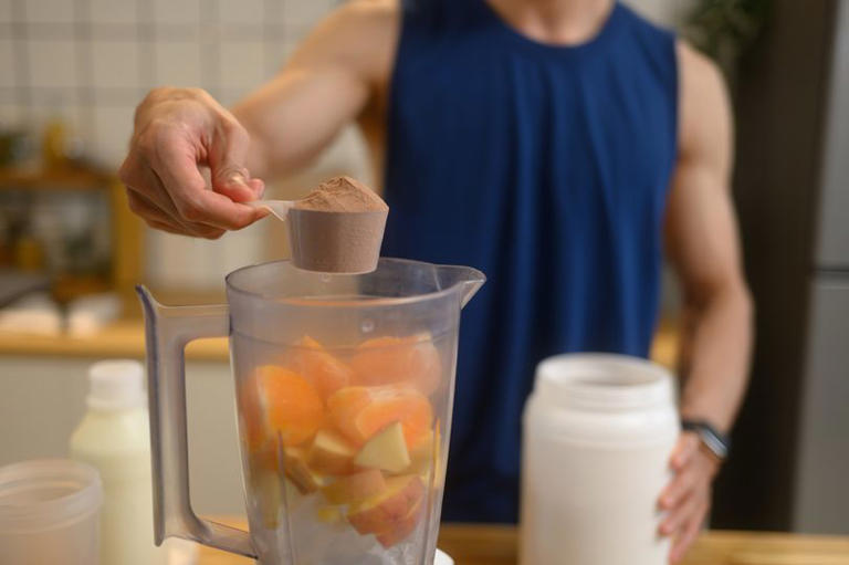 Man adding a scoop of chocolate protein powder into a blender filled with chopped fruits, preparing nutritious shake in the kitchen.